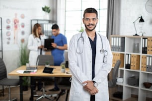 Young 30-aged handsome pleasant Arabic male doctor in scrubs and white coat, posing at camera indoors at modern light office in clinic. Blurred view of two multiethnic colleagues talking on background