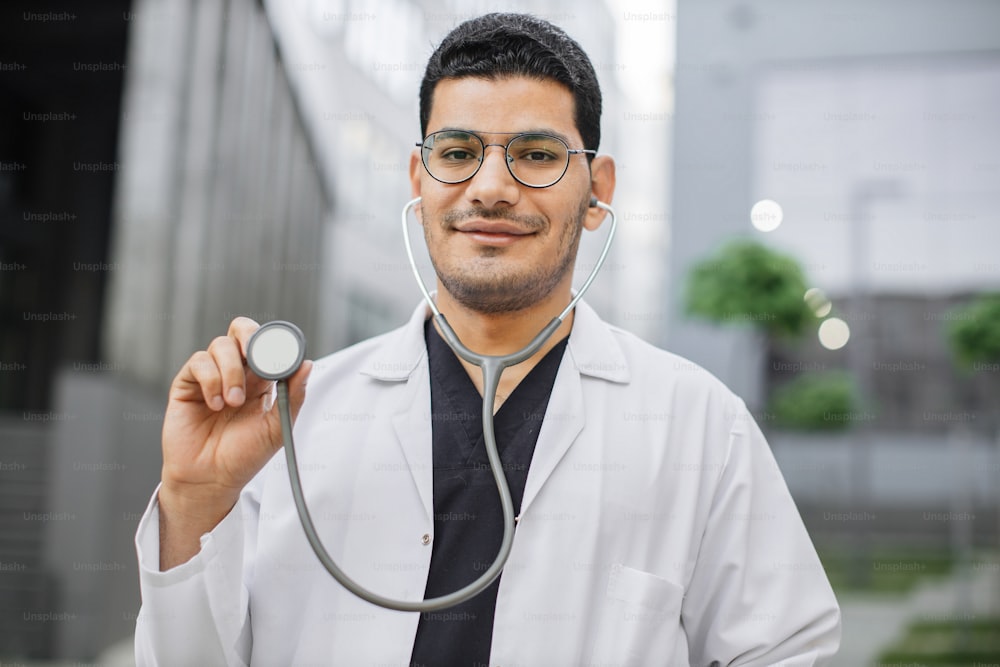 Portrait of smiling friendly male Asian doctor or medical student in eyeglasses, standing outside modern hospital, demonstrating his stethoscope to camera. Focus on face