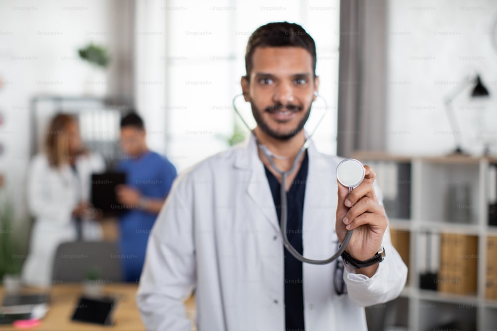 Close up blurred view of handsome male Indian doctor, standing with stethoscope in modern light hospital boarding room with two diverse colleagues talking on the background. Focus on stethoscope