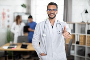 Medical education and healthcare. Blur view with copy space of young smiling male medical student showing thumb up to camera while standing in modern study room with groupmates on background