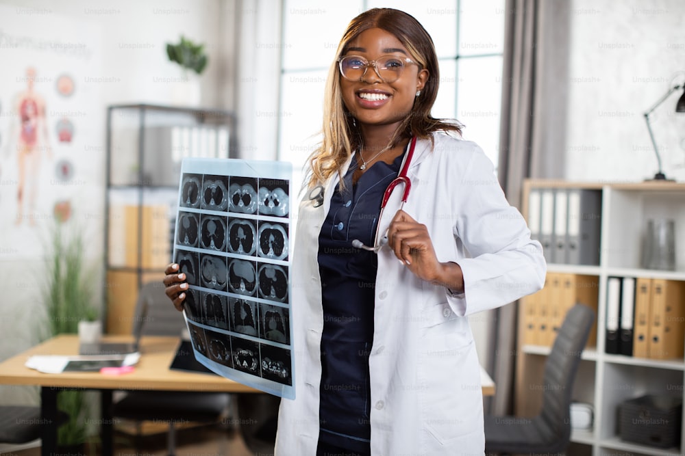 Medical equipment, radiology, people and occupation concept. Portrait of pretty smiling skilled black skinned woman gp doctor, posing to camera in her modern private office with MRI scan in hands