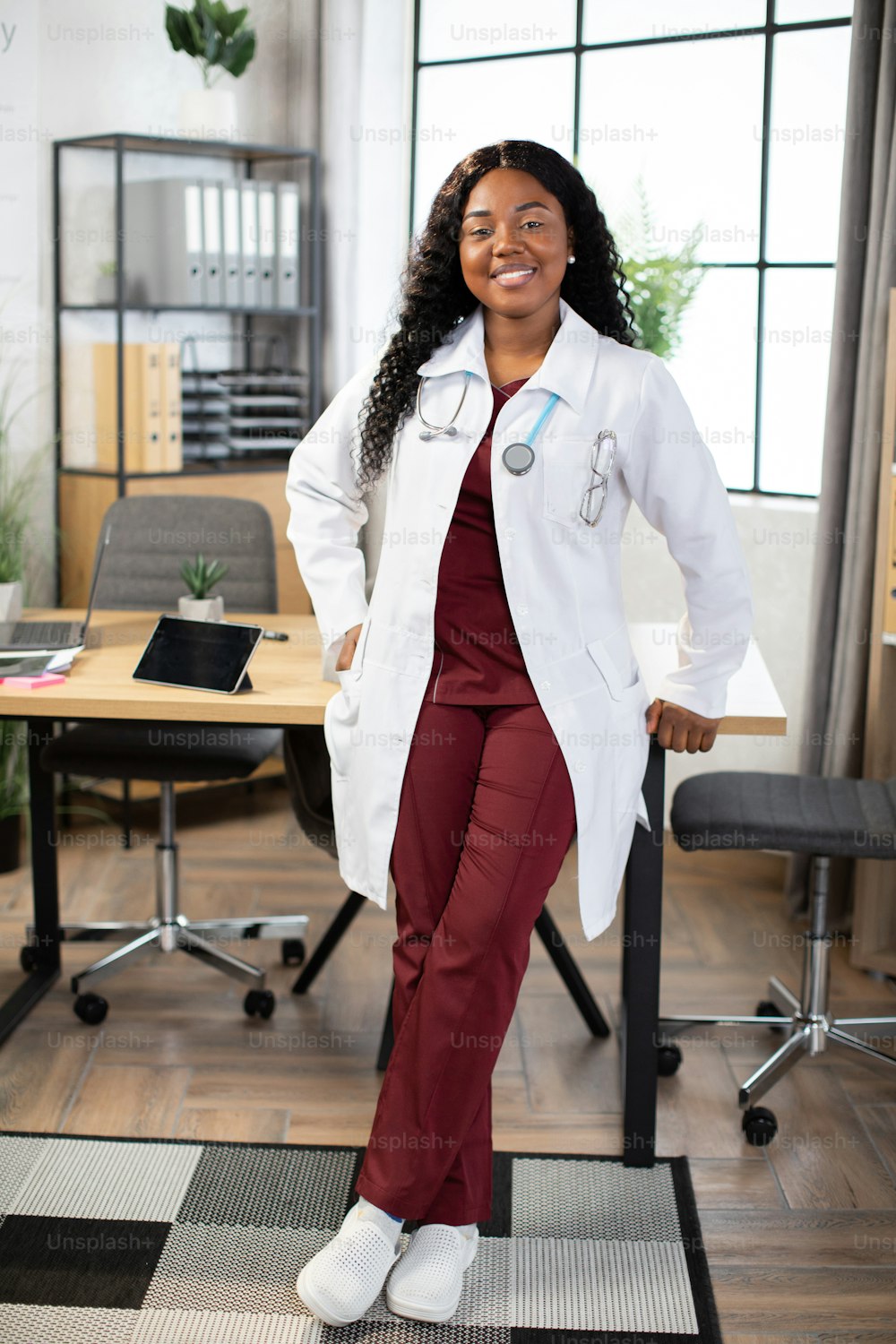 Full-length portrait of friendly 30-aged pretty Afro-American woman doctor, posing to camera with happy smile, while leaning on the table in her modern hospital office