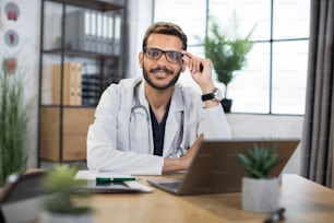 Nahaufnahme eines lächelnden, gutaussehenden männlichen malaiischen Arztes im Alter von 30 Jahren mit Brille, der mit Laptop in seinem Privatbüro am Tisch sitzt und in die Kamera schaut.