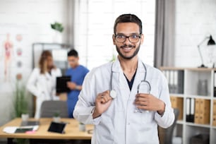Bonito e simpático jovem médico árabe malaio do sexo masculino está olhando para a câmera e sorrindo enquanto seus colegas estão de pé e falando ao fundo