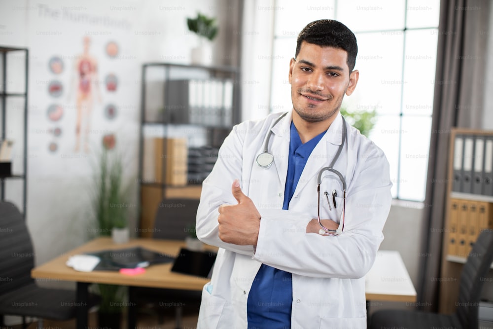 Closeup portrait of a happy young handsome Arabian Muslim physician, with stethoscope around neck, posing with arms crossed and showing thumb up to camera, standing in modern office. Copy space