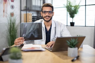 Making a diagnosis concept. Portrait of young qualified male Asian Indian doctor, sitting in office, holding skull xray image, working on laptop computer, smiling at camera