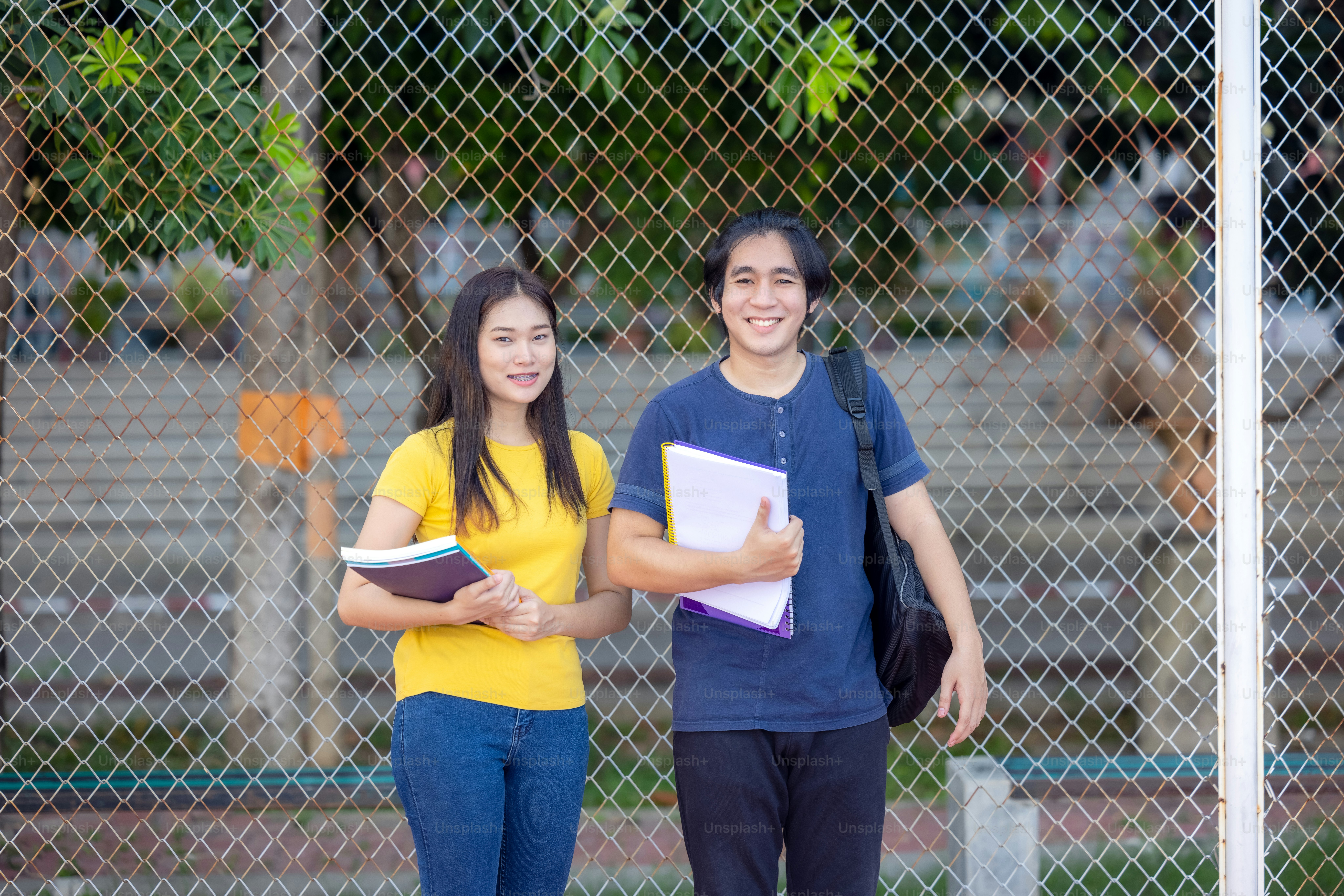 Outside of school, a happy young couple of students stands along a fence, studying a book