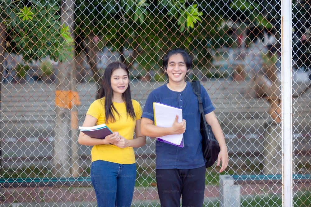 Outside of school, a happy young couple of students stands along a fence, studying a book.