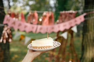 Pedazo de pastel de cumpleaños moderno con vela en la mano de la mujer sobre fondo de guirnalda rosa de feliz cumpleaños en el parque. Celebrando el cumpleaños en una fiesta de picnic al aire libre. Pide un deseo