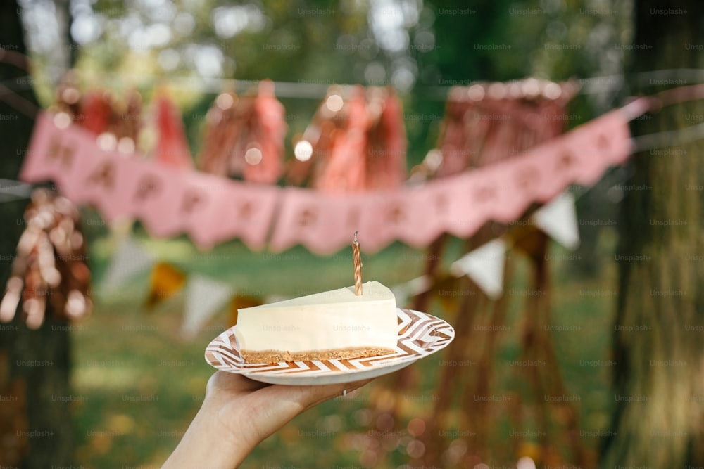 Piece of modern birthday cake with candle in woman hand on background of pink happy birthday garland in park. Celebrating birthday at picnic party outdoor. Make a wish