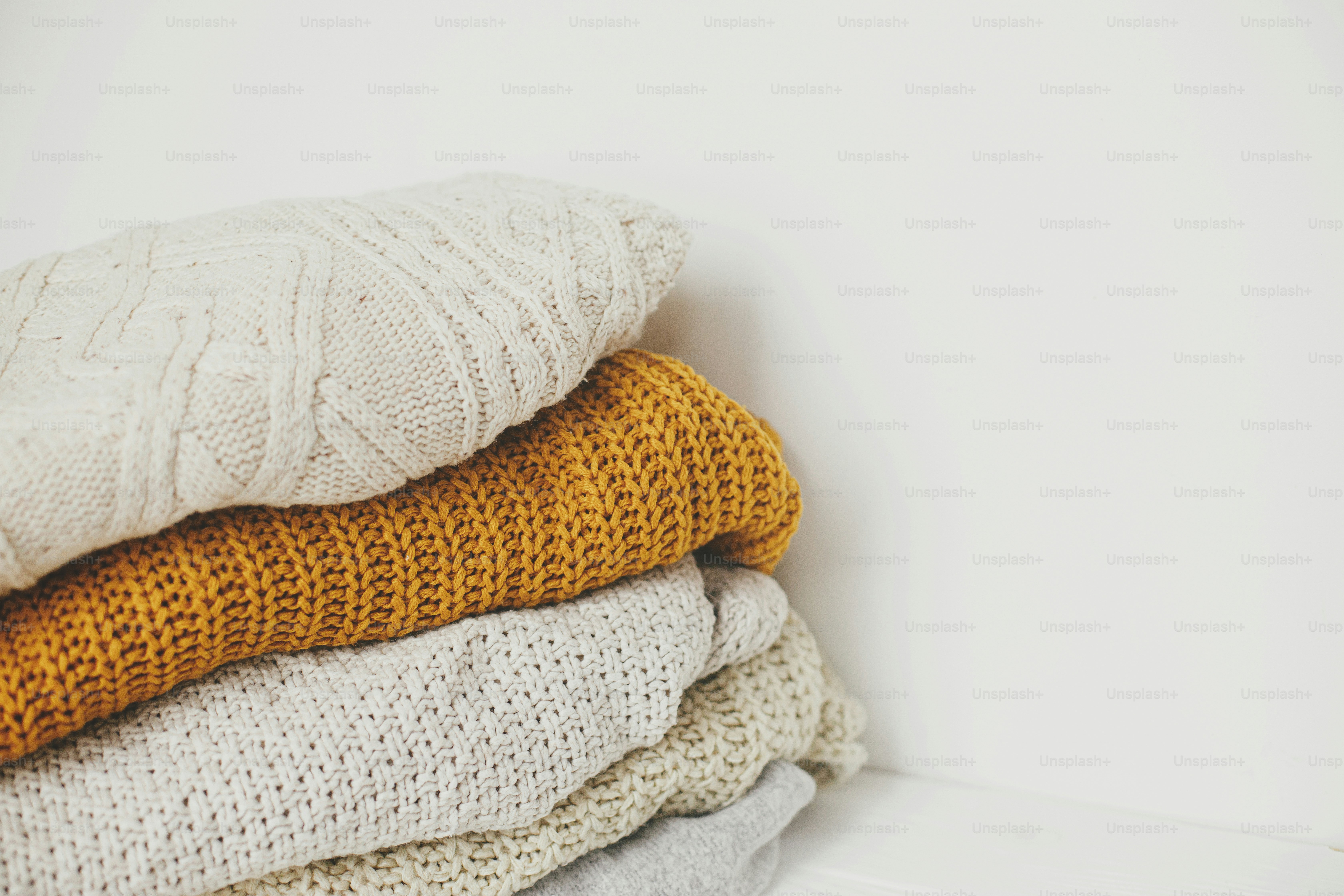 Pile of white, yellow and beige knit sweaters close up in white scandinavian room. Cozy knitted sweaters. Hello autumn. Stylish wardrobe for cold season. Space for text