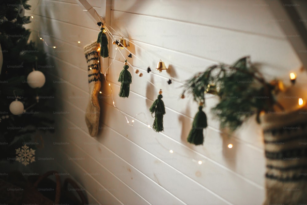 Christmas rustic stocking with fir branch and stylish garland with bells and trees on white wooden wall with christmas lights. Festive decorated atmospheric scandinavian room. Boho handmade decor