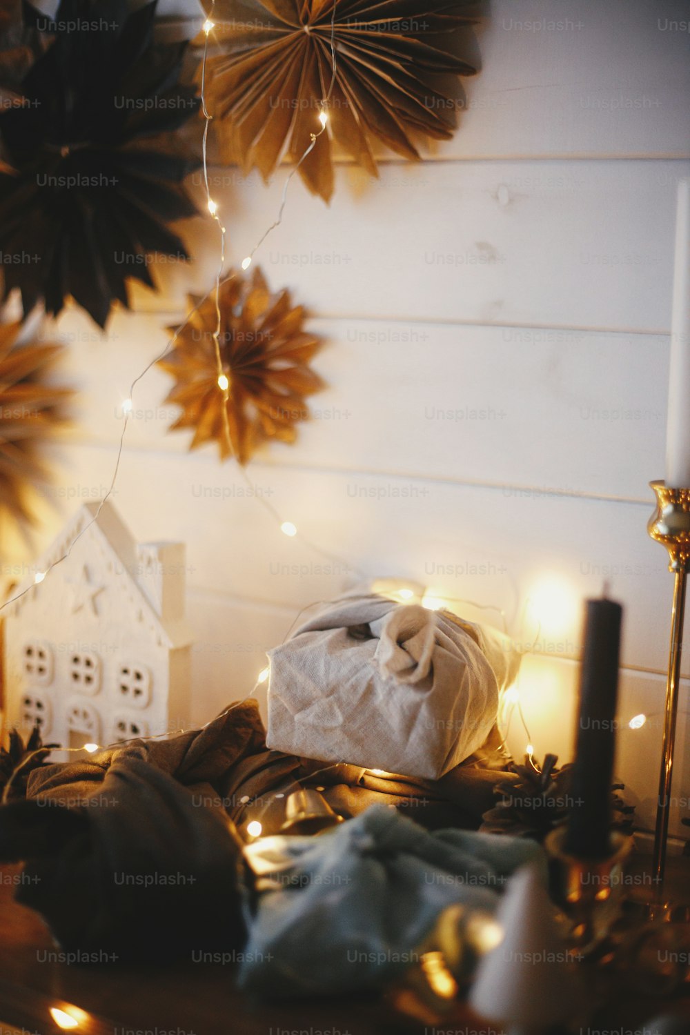 Buon Natale! Eleganti regali di Natale senza plastica su scaffale di legno sullo sfondo di casetta, alberi, stelle di carta e bokeh di luci dorate natalizie. Suggestiva sala scandinava festiva