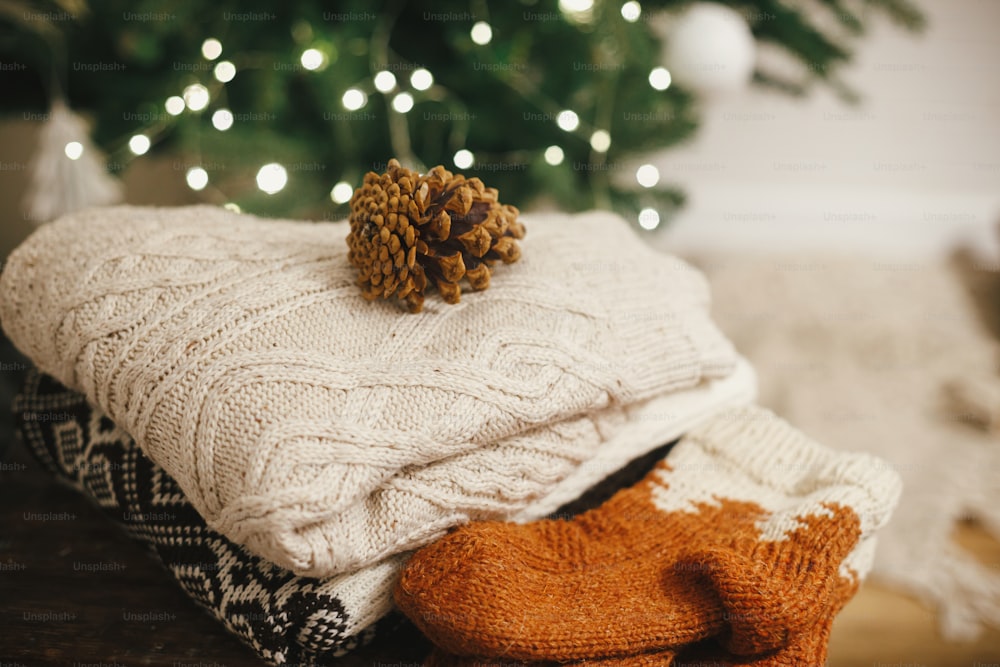 아늑한 니트 스웨터와 양모 양말은 크리스마스 트리 조명을 배경으로 솔방울이 있는 소박한 나무에 있습니다. 축제 장식 스칸디나비아 방에 세련된 겨울 천 더미. 텍스트를 위한 공간