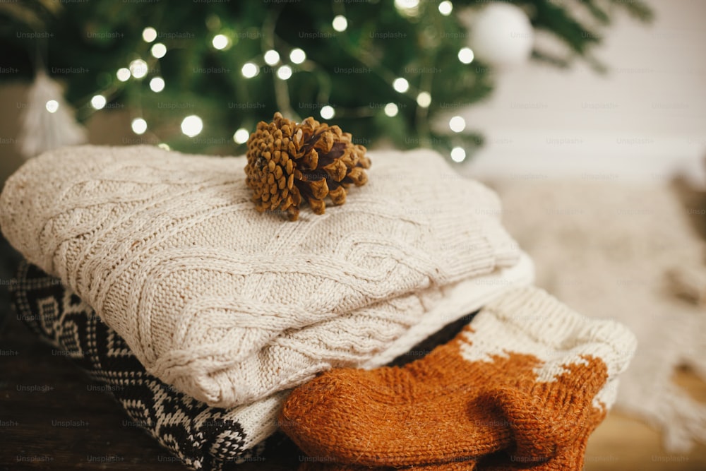 Cozy knitted sweaters and wool socks on rustic wood with pine cone on background of christmas tree lights. Pile of stylish winter cloth in festive decorated scandinavian room. Space for text