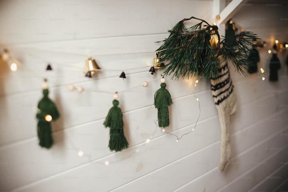 Weihnachtslicher rustikaler Strumpf mit Tannenzweig und stilvoller Girlande mit Glocken und Bäumen an weißer Holzwand mit Weihnachtsbeleuchtung. Festlich dekoriertes stimmungsvolles skandinavisches Zimmer. Modernes handgefertigtes Dekor