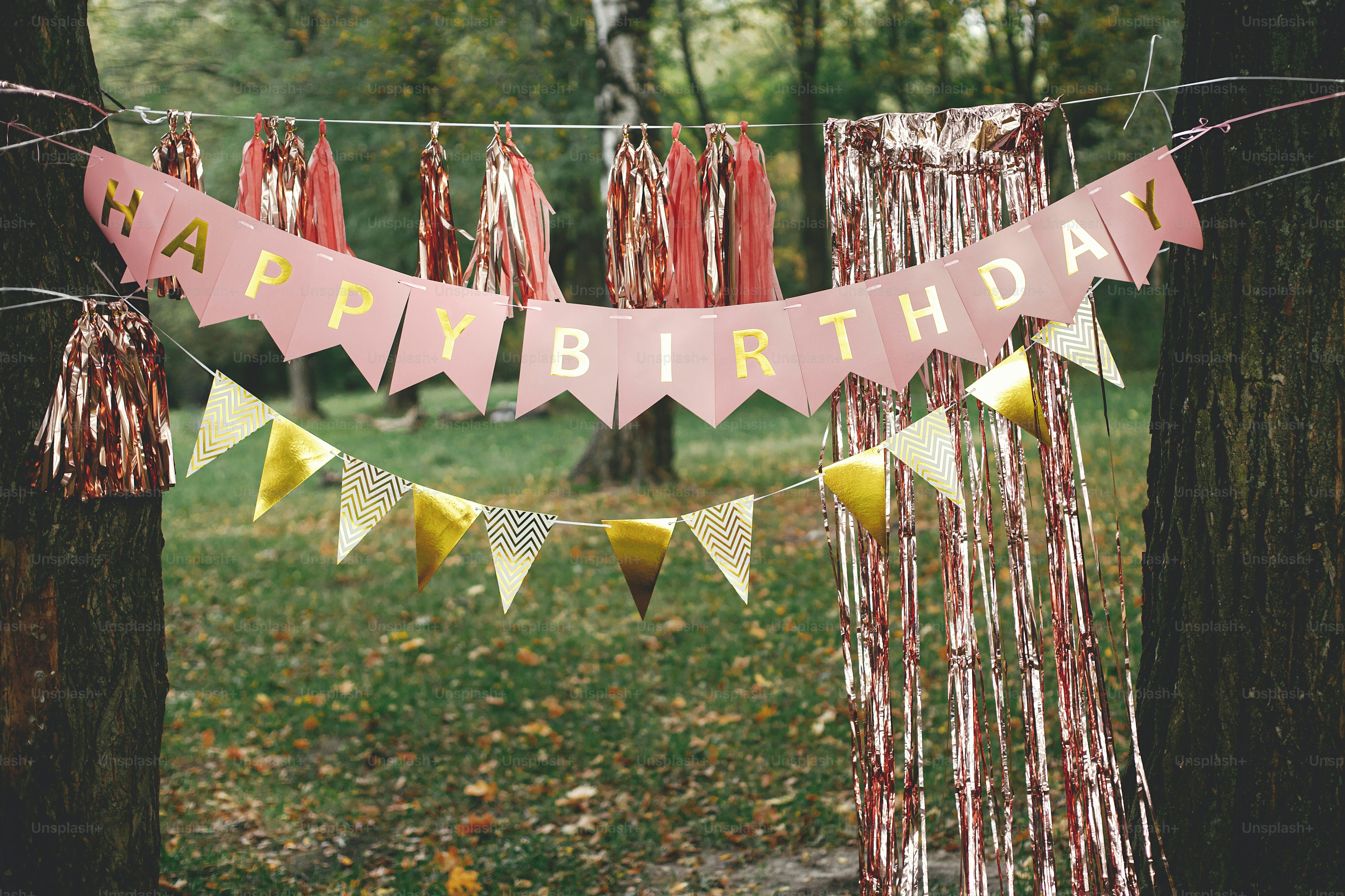 Stilvolle Geburtstagsgirlande im Park. Modernes Roségold-Dekor, Quastengirlande und Happy Birthday-Banner hängen an Bäumen Picknick-Party. Geburtstagsfeier im Freien