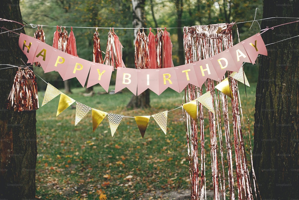 Stylish happy birthday garland hanging in park. Modern rose gold decor, tassel garland and happy birthday banner hanging on trees picnic party. Birthday celebration outdoor