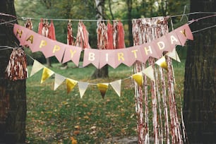 Stilvolle Geburtstagsgirlande im Park. Modernes Roségold-Dekor, Quastengirlande und Happy Birthday-Banner hängen an Bäumen Picknick-Party. Geburtstagsfeier im Freien