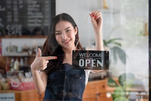 Portrait d’une jeune fille de barista souriante en tablier tenant un panneau ouvert tout en se tenant debout dans son café. Le personnel féminin élégant d’un café asiatique tourne la plaque de porte le matin dans sa propre petite entreprise de magasin.
