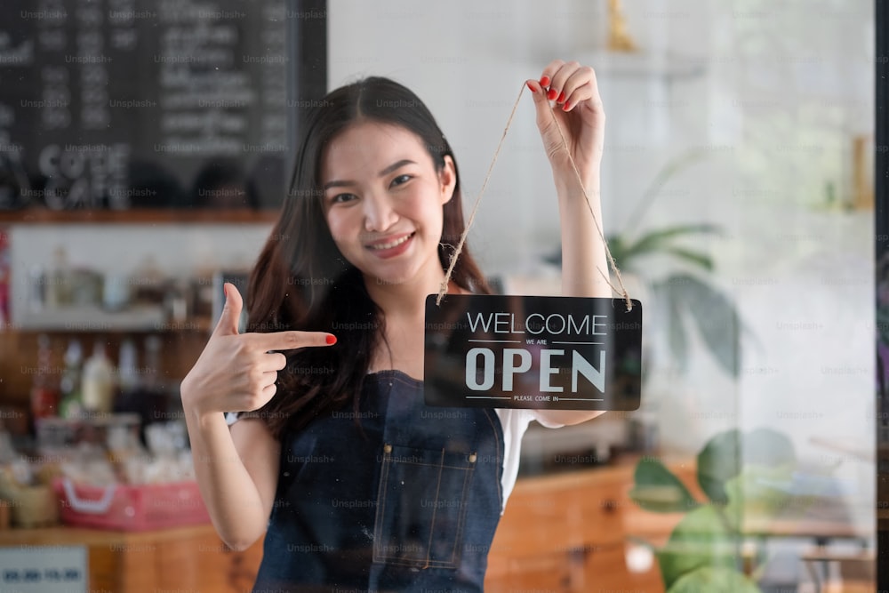 Porträt eines lächelnden jungen Barista-Mädchens in Schürze, das ein offenes Schild hält, während es in ihrem Café steht. Elegantes asiatisches Café weibliche Mitarbeiter drehen Türplatte am Morgen in eigenem Laden kleines Geschäft.