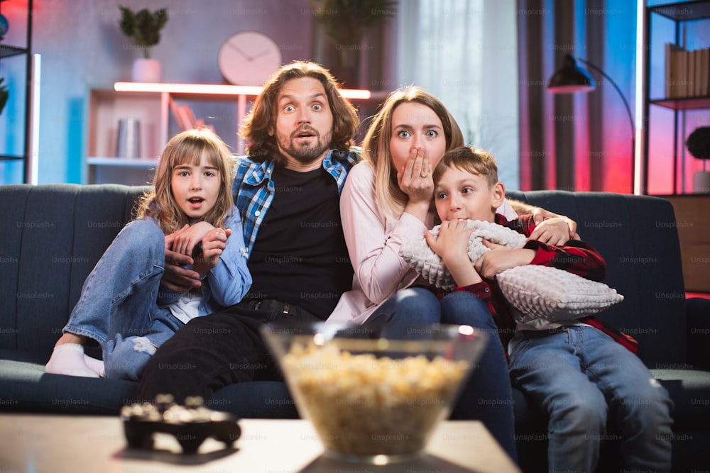 Padres jóvenes asustados o sorprendidos con un hijo y una hija pequeños viendo una película de terror impresionante en la televisión durante la noche en casa. Emociones humanas reales. Concepto de ocio familiar.