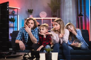 Unhappy Caucasian family of four in casual wear sitting on couch and feeling bored while watching television. Concept of people, house and television.