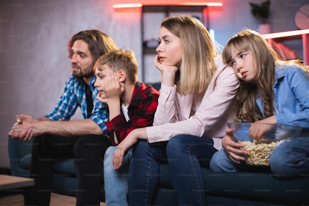 Young parents with two kids sitting together on couch and feeling tired of boring movies on TV. Domestic life of caucasian family.