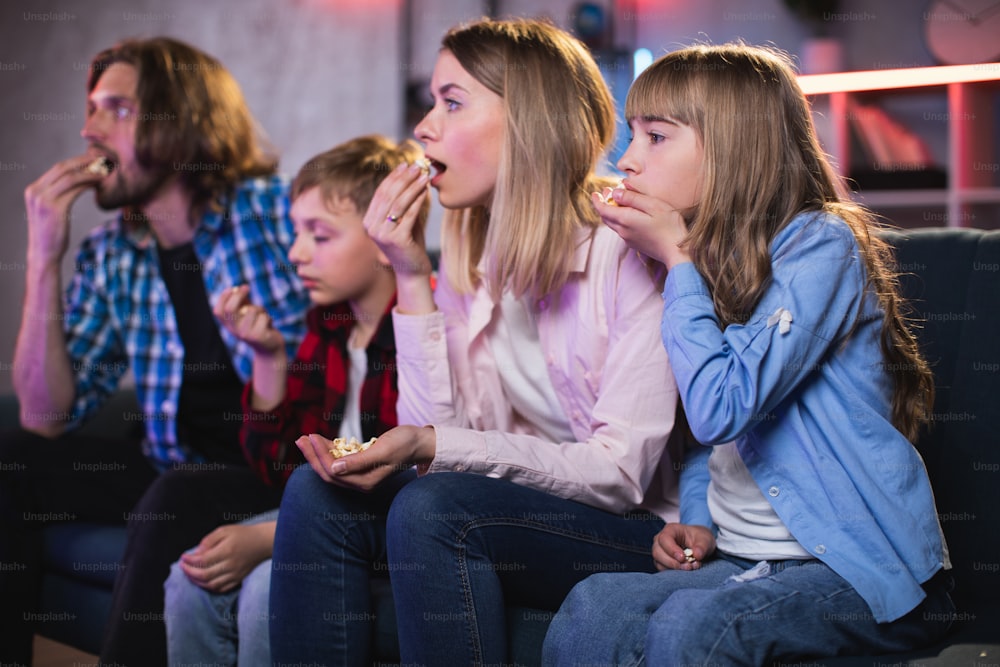 Vista laterale di genitori stupiti con due bambini che guardano la TV e mangiano popcorn durante la sera. Giovane famiglia seduta insieme sul divano e sentimento di eccitazione.