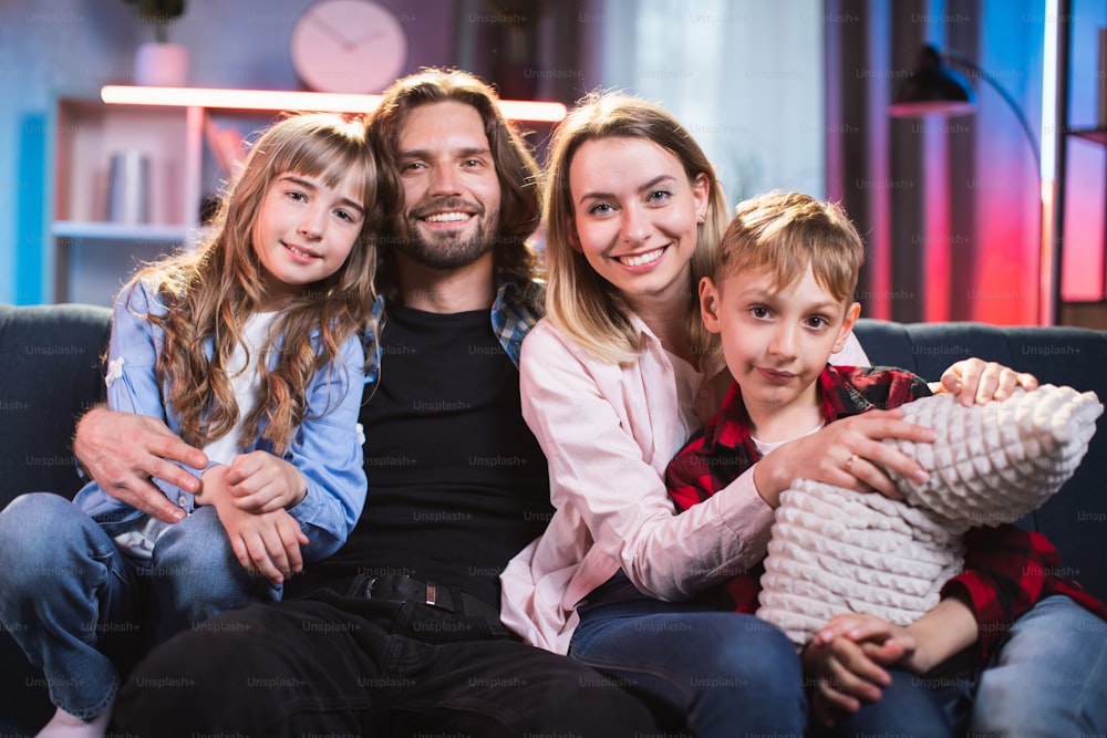 Hermosa familia positiva con ropa informal sentada en abrazos en un cómodo sofá y pasando la noche para ver la televisión. Padres con niños juntos en casa.
