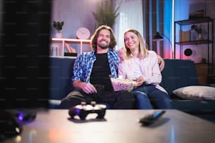 Positive young couple watching movie on TV and eating popcorn at home. Happy caucasian man and woman sitting on couch and hugging. Time together.