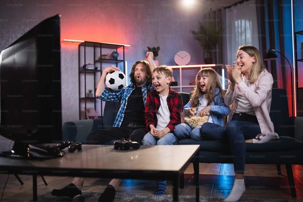 Emotional parents with little son and daughter celebrating goal of favorite team during soccer match. Satisfied family sitting on couch and watching sport competition on TV.