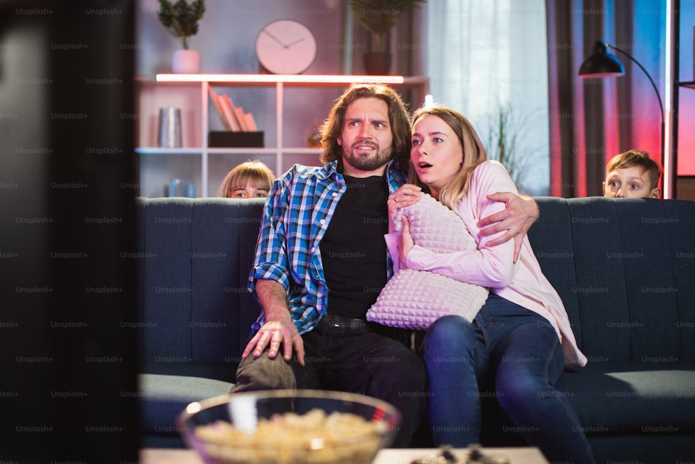 Homem caucasiano abraçando sua mulher aterrorizada enquanto assiste a um filme de terror em casa. Duas crianças escondidas atrás do sofá. Emoções humanas reais.