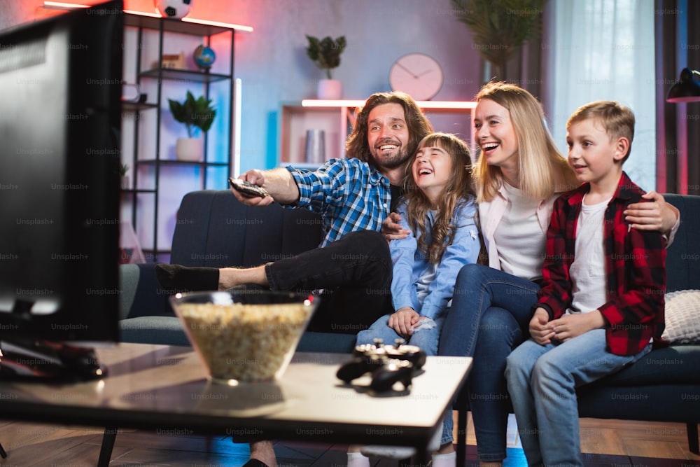 Cheerful man and woman with two children sitting in hugs on comfy sofa and using remote control while watching television. Happy family with real emotions on faces at home.