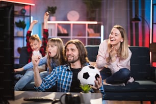 Padres jóvenes positivos con un hijo y una hija pequeños sentados en el sofá y viendo partidos de fútbol en la televisión. Tiempo libre de la familia en casa.