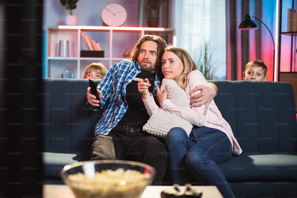 Marido barbudo assustado abraçando sua esposa assustada durante, assistindo a filmes de terror na TV em casa à noite. Duas crianças assustadas estão escondidas do susto atrás do sofá