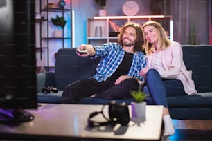 Beautiful caucasian couple sitting in hugs on comfy couch and watching TV. Smiling wife and husband with remote control choosing evening movie at home.