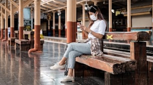 Adolescente turista asiática usando máscara esperando estação de trem de distância usando smartphone para mapa on-line, check-in de mídia social ou reserva de bilhetes. Tecnologia moderna de aplicativos de viagem, viajante solitário, conceito de aventura ferroviária de férias de verão