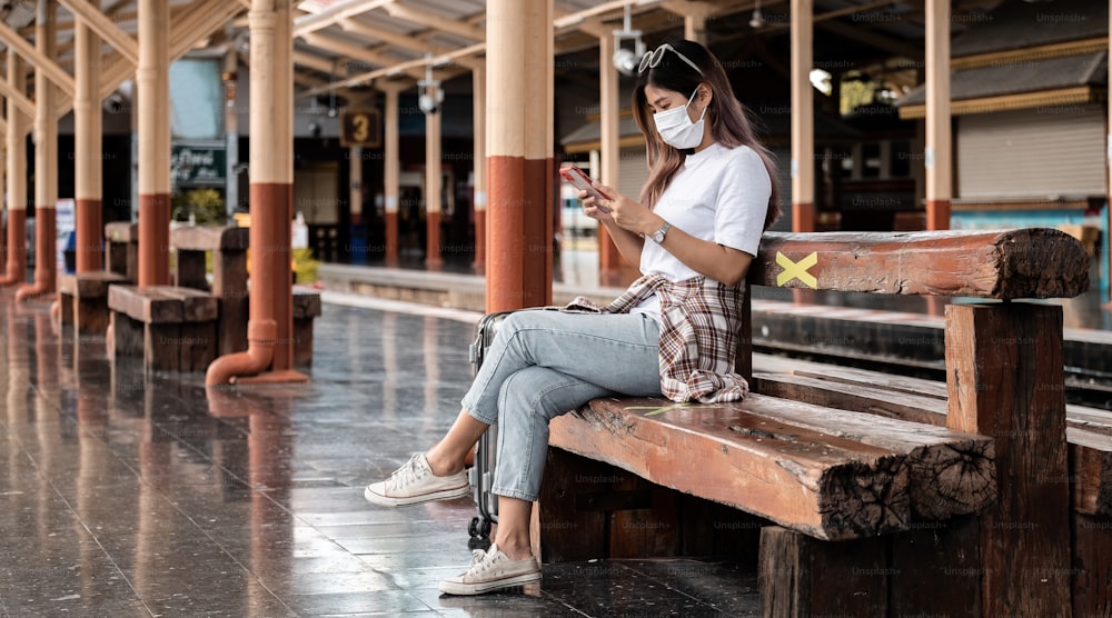 Ragazza adolescente turista asiatica che indossa la maschera aspettando la stazione ferroviaria utilizzando lo smartphone per la mappa online, il check-in sui social media o la prenotazione del biglietto. Moderna tecnologia dell'app di viaggio, viaggiatore solitario, concetto di avventura ferroviaria per le vacanze estive