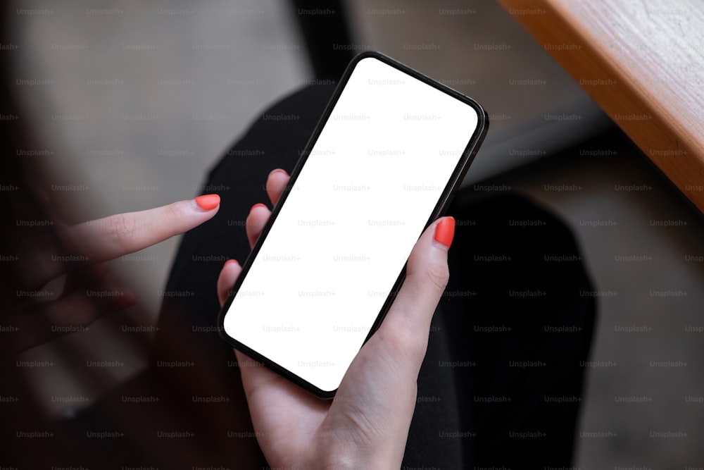 카페에 앉아 있는 동안 빈 흰색 화면이 있는 검은색 휴대폰을 들고 있는 여성의 상위 뷰 모형 이미지