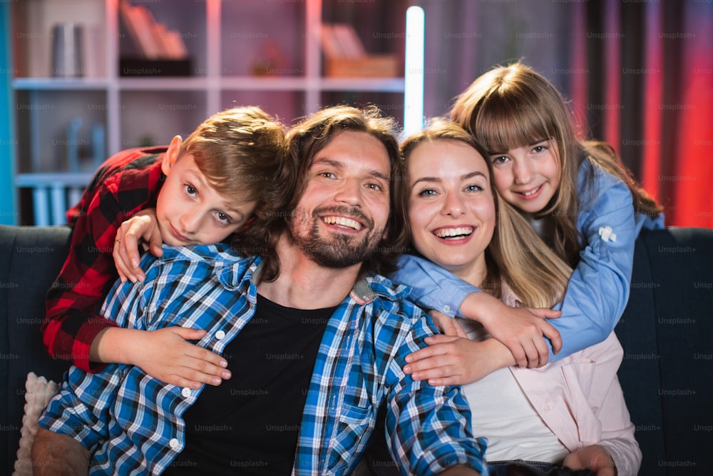 Portrait of beautiful and happy family in casual wear resting together on couch during evening time. Pretty kids hugging lovely parents. Idyllic atmosphere at home.