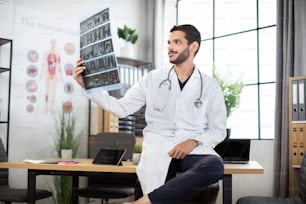 Close up of young confident Asian Indian male physician looking at computer tomography x-ray image, while working in modern bright medical office. MRI scan and diagnostics concept