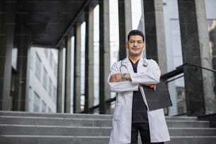 Abile medico maschio indù arabo fiducioso in camice bianco e scrub, con stetoscopio, tenendo appunti, in piedi con le braccia incrociate sullo sfondo della clinica moderna. Copiare lo spazio per il testo