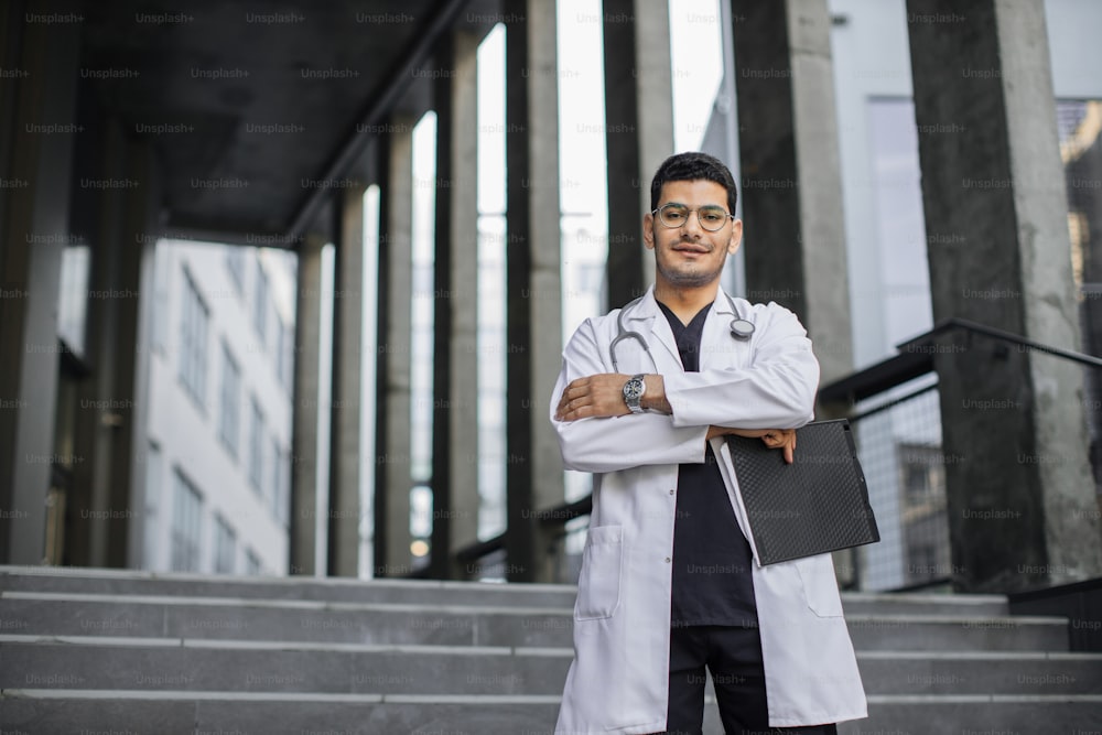 Skilled confident Arabian hindu male physician in white coat and scrubs, with stethoscope, holding clipboard, standing with arms crossed on the background of modern clinic. Copy space for text