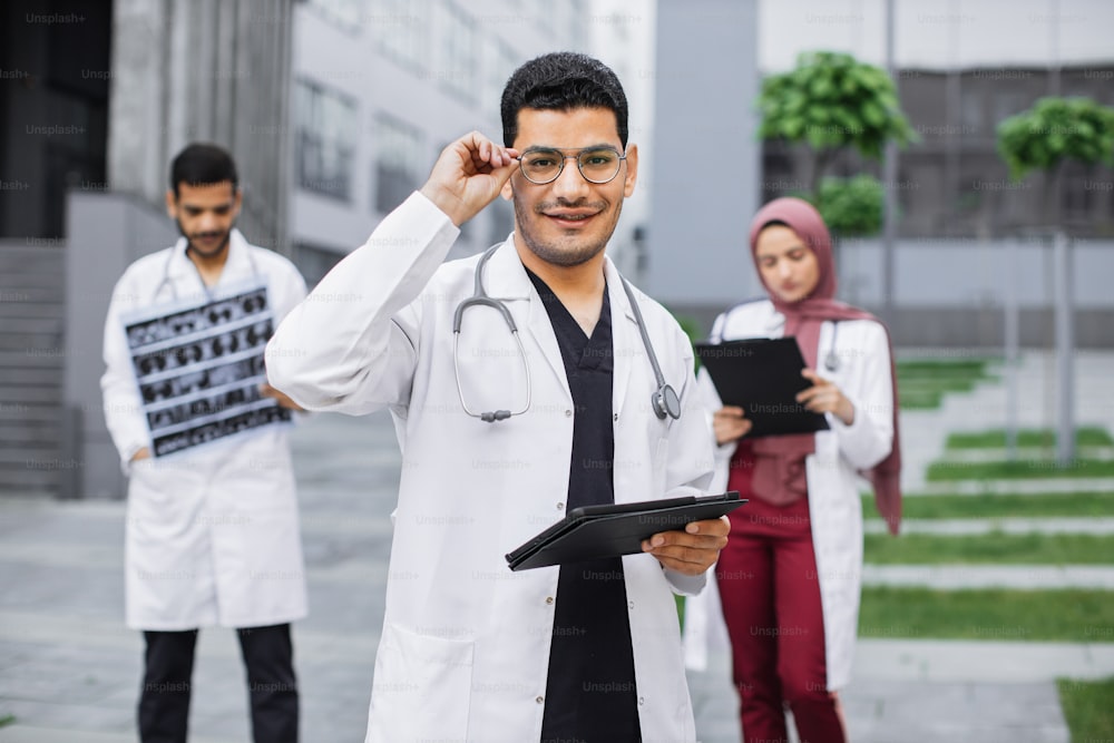 Coworking all'aperto dell'ospedale. Giovane medico arabo indù in occhiali utilizzando il dispositivo tablet, in posa per la fotocamera con sorriso. Medici e medici multietnici che lavorano sullo sfondo