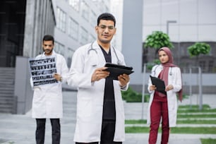 Three young doctors wearing white coat, working with tablet pc, xray and patient's medical record outside the hospital. Male hindu doctor typing on tablet computer, standing in front of colleagues