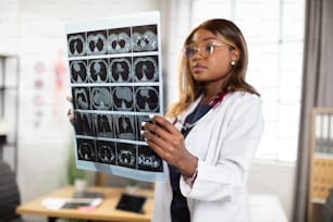 Close up of young confident afro-american female physician looking at computer tomography x-ray image, while standing in modern bright medical office. Selective focus on MRI scan