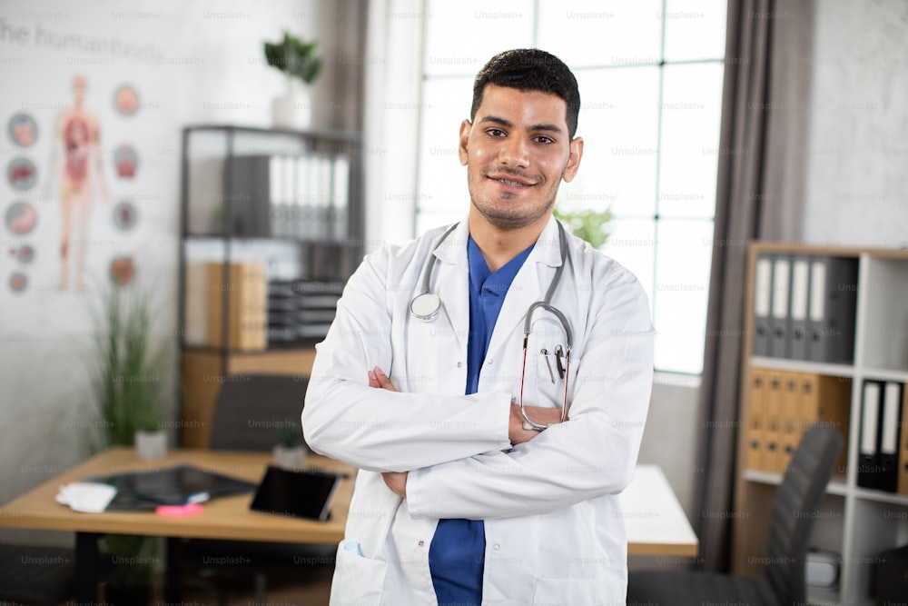 Smiling young 30-aged male Arabic Asian doctor posing with arms crossed in the office. Male physician in blue suit and white coat, with a stethoscope around neck, posing to camera indoors