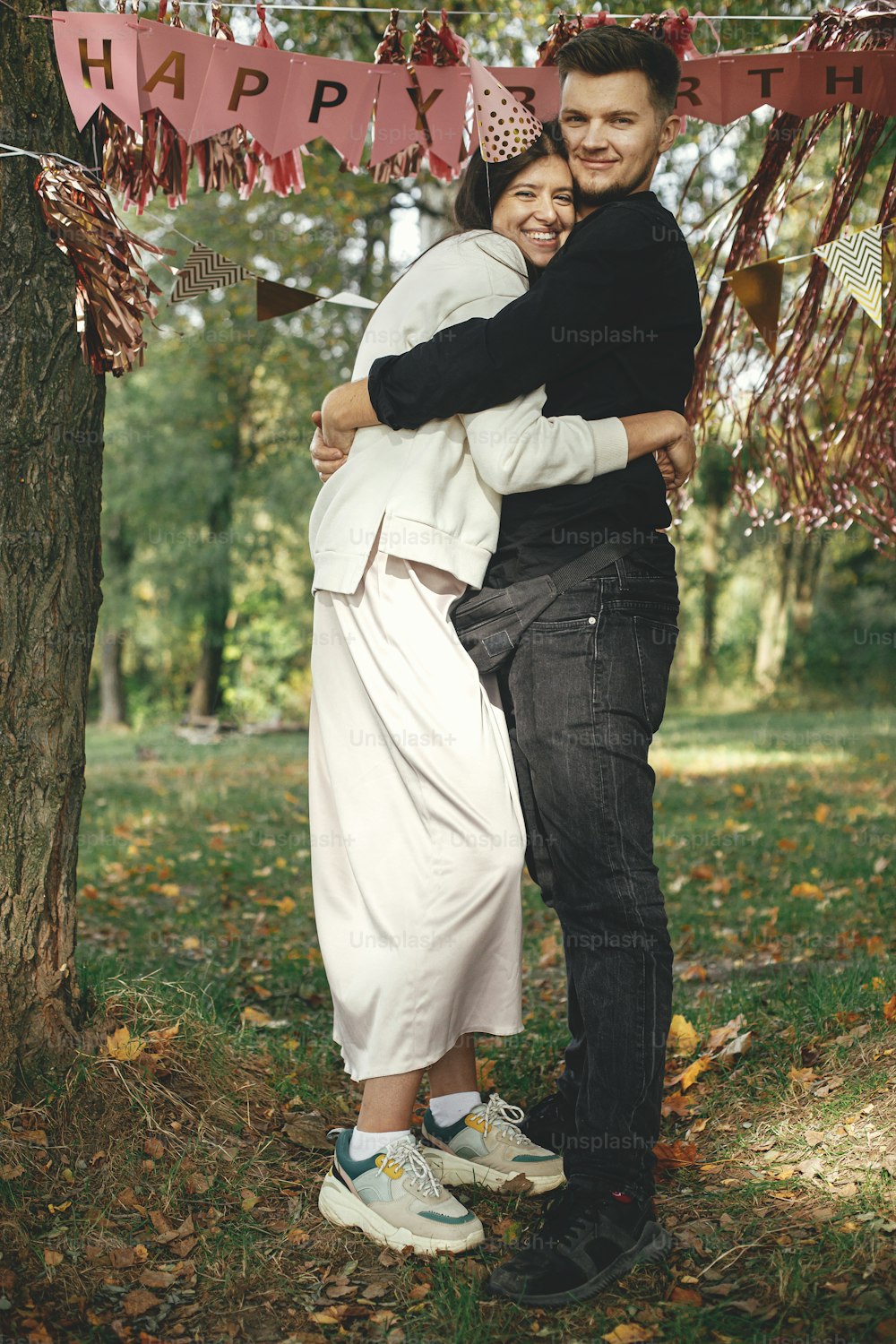 Young stylish couple celebrating birthday with family and friends at picnic party outdoor. Happy woman in party hat and handsome man hugging on background of pink happy birthday garland in park