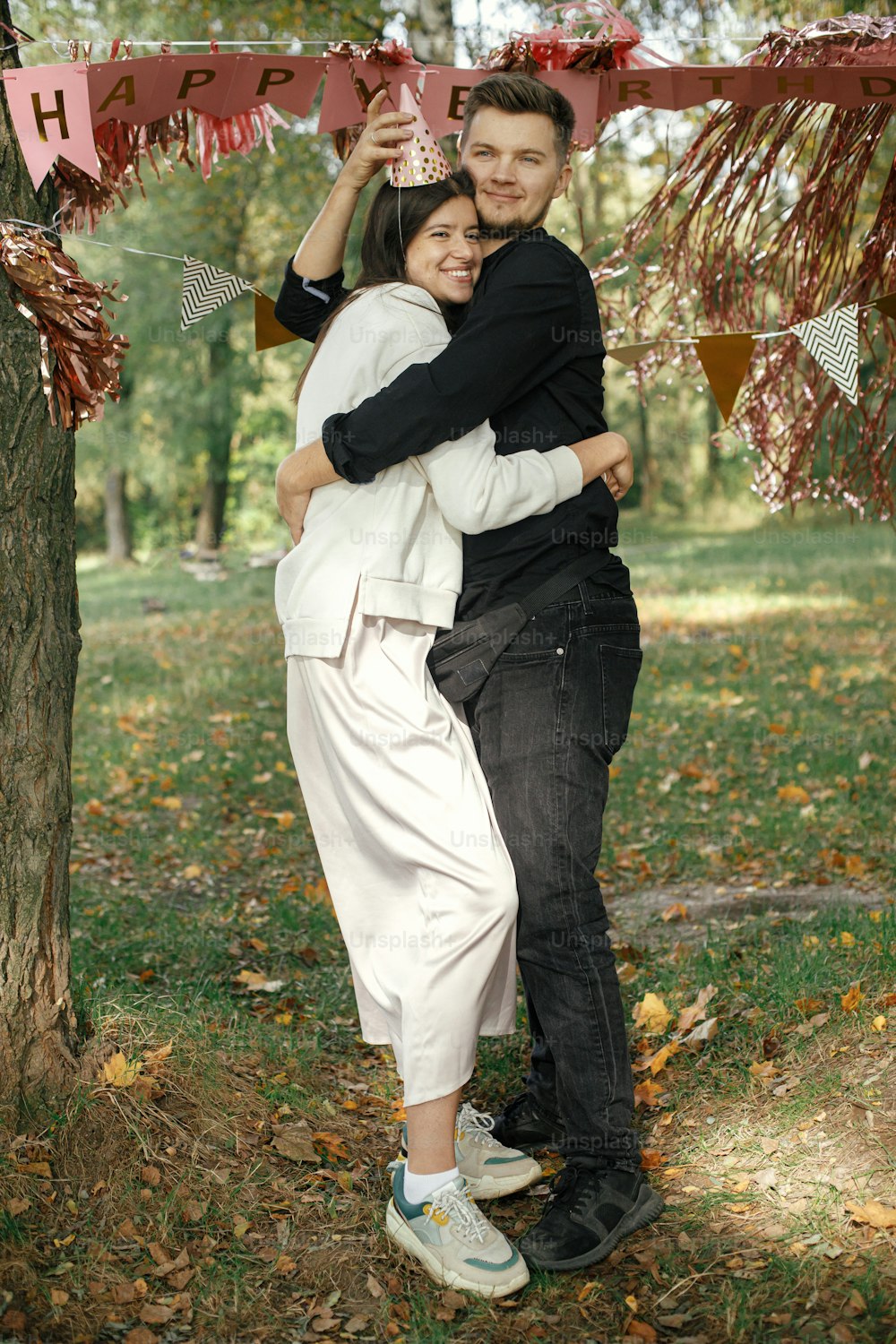 Young stylish couple celebrating birthday with family and friends at picnic party outdoor. Happy woman in party hat and handsome man hugging on background of pink happy birthday garland in park